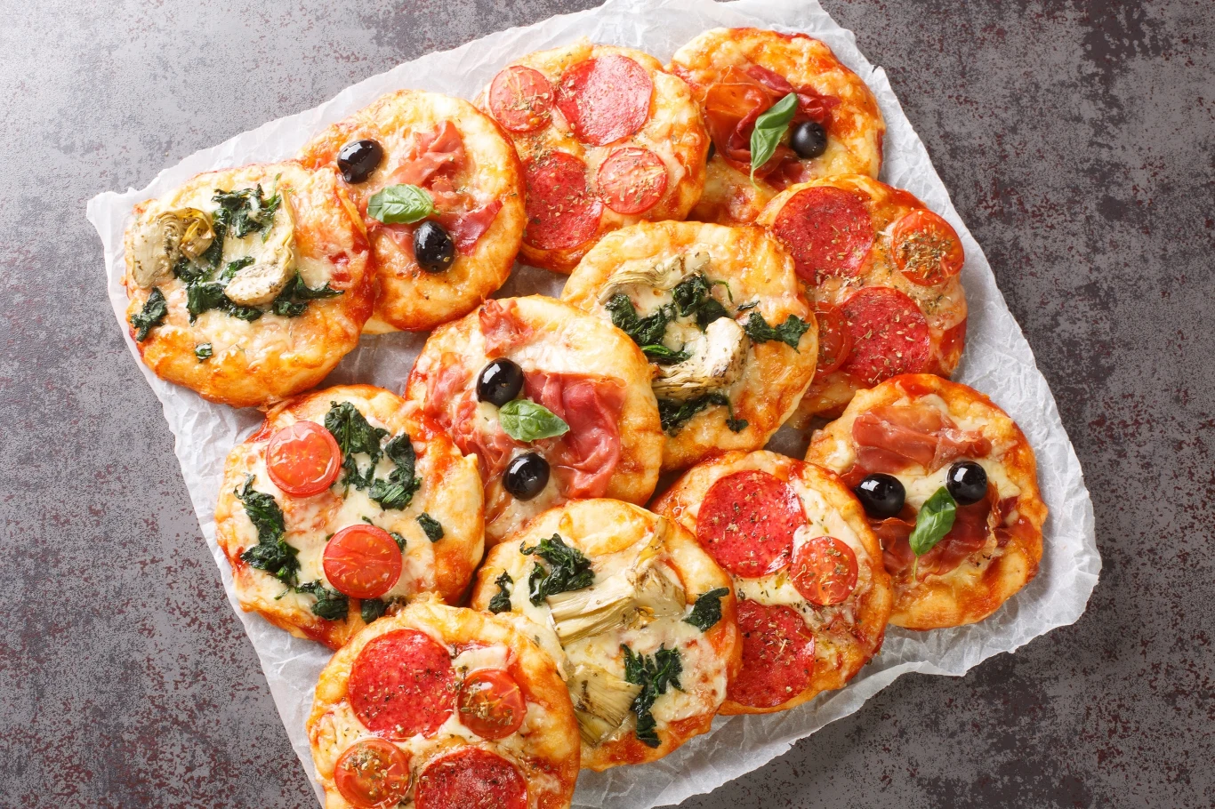 Poznaj przepis na pizzerinki z serka wiejskiego. To idealne przekąski czy pomysł na ciepłą kolację. Pizzerinki z różnymi dodatkami, w tym pomidorami, szynką, serem i bazylią, ułożone razem na papierze do pieczenia.