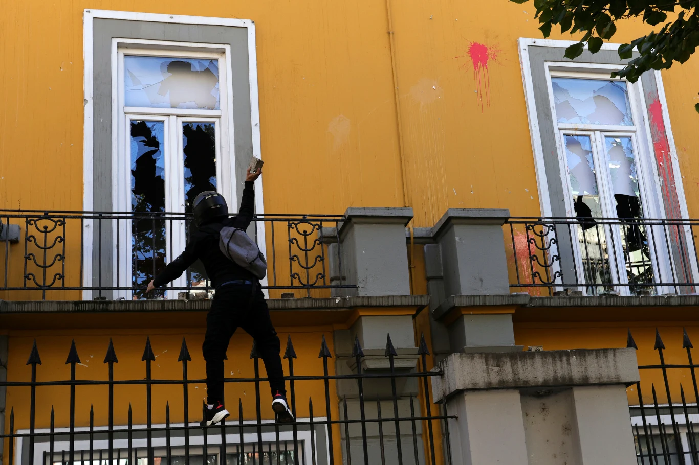 Zamaskowany demonstrant rzuca kamieniem w kierunku budynku z wybitymi szybami podczas protestów w Tiranie.