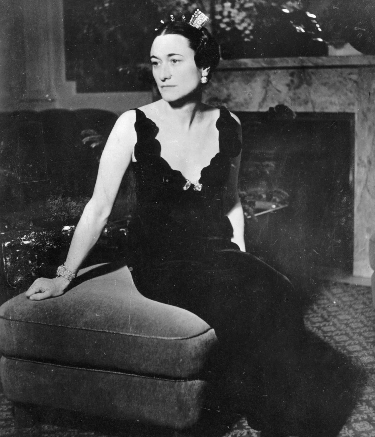 Wallis Simpson, 1 stycznia 1937 Kobieta w eleganckiej czarnej sukni z głębokim dekoltem, siedząca na pufie, z ozdobami we włosach i biżuterią, w wyraźnie zamyślonym nastroju.