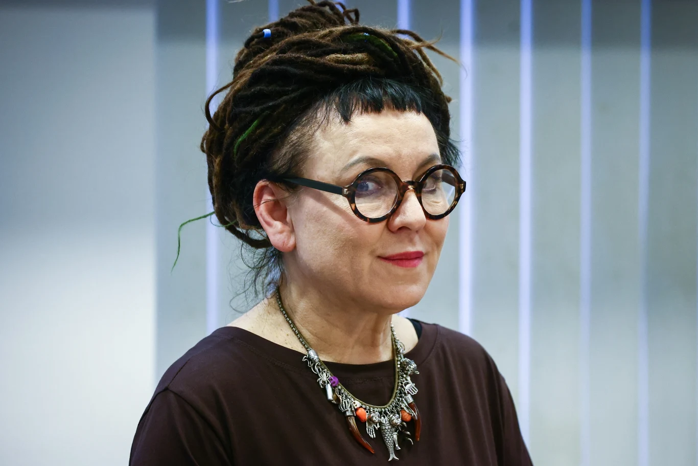 Olga Tokarczuk w Gliwicach. Jak zdobyć wejściówkę na spotkanie? Olga Tokarczuk w ciemnych okularach i naszyjniku, stoi na tle jasnych pionowych żaluzji, kontekst spotkania w Gliwicach.
