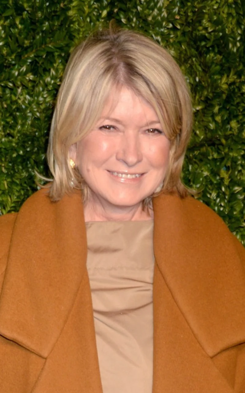 Martha Stewart jest gwiazdą w Stanach Zjednoczonych, ale ma polskie korzenie Kobieta z blond włosami, ubrana w elegancki płaszcz w odcieniu karmelowym oraz beżową bluzkę, stoi na tle zielonej ściany z roślin.