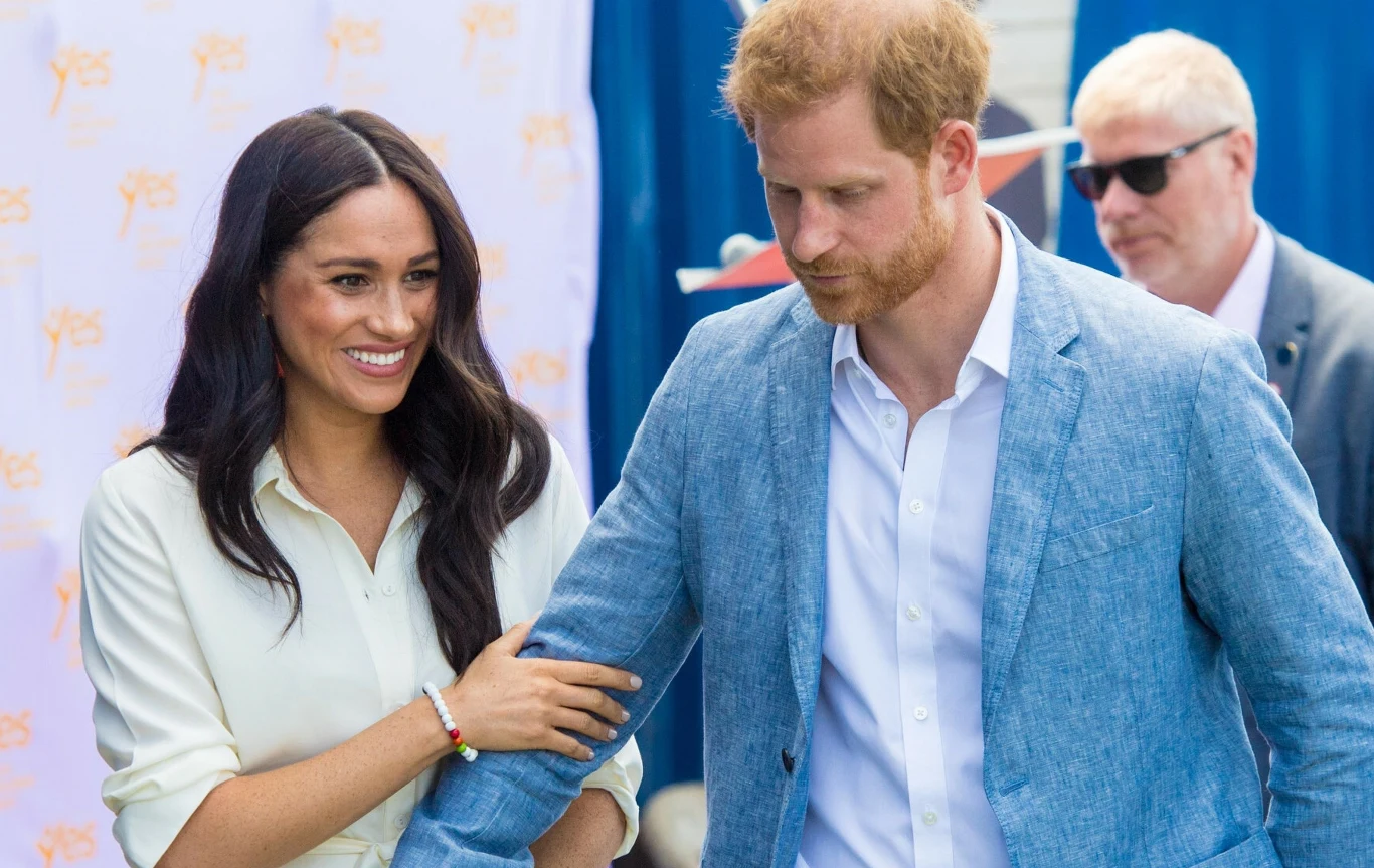 Meghan Markle i książę Harry Meghan Markle i książę Harry podczas wspólnego wystąpienia. Obok nich stoi mężczyzna w okularach przeciwsłonecznych.