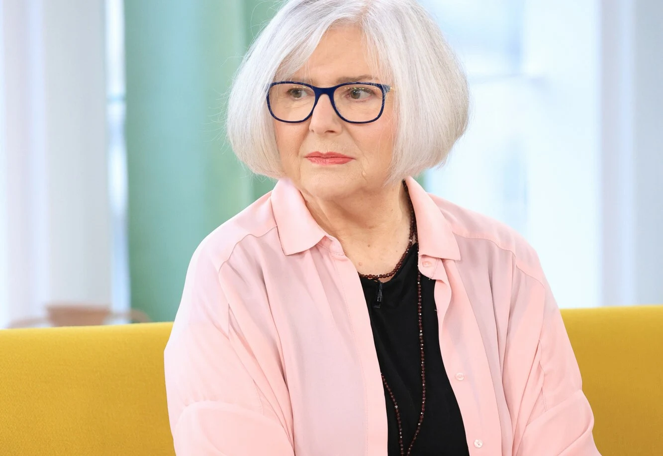 Grażyna Bukowska Starsza kobieta z siwymi włosami i okularami w ciemnych oprawkach, ubrana w jasnoróżową koszulę i czarną bluzkę, siedzi na żółtej sofie.