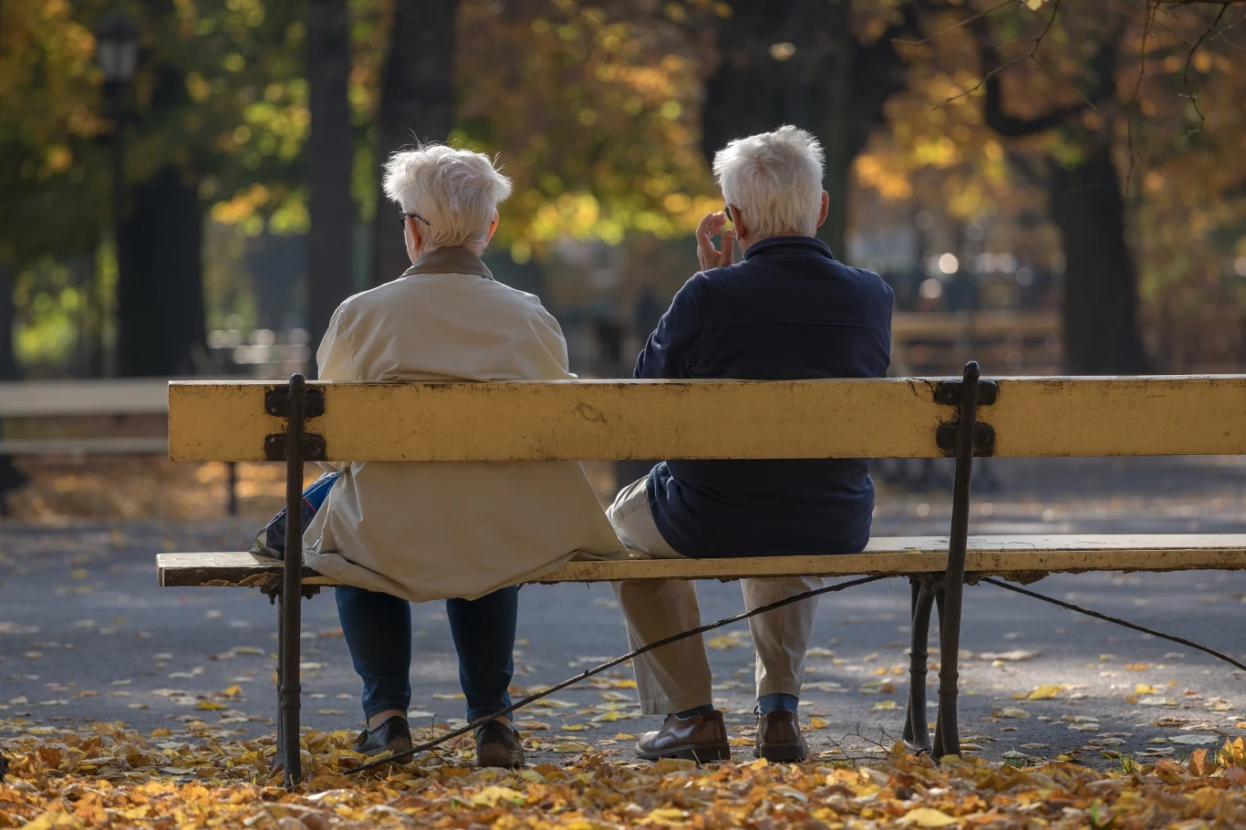 Seniorzy z Ełku mogą liczyć na dodatkową pomoc. Rusza kolejna edycja miejskiego programu. Dwoje starszych osób z siwymi włosami siedzi na ławce w jesiennym parku i rozmawia, kontekst programu w Ełku.