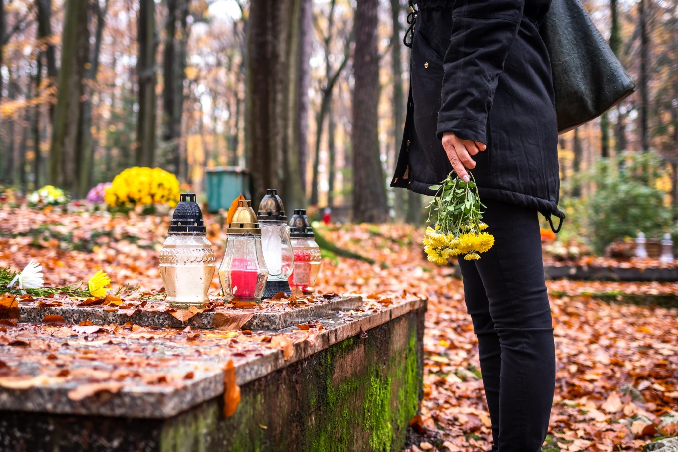 Przesąd na Wszystkich Świętych. Tego lepiej nie robić przed wizytą na cmentarzu. Osoba w ciemnych ubraniach z żółtymi kwiatami przy grobie ze zniczami i jesiennymi liśćmi.