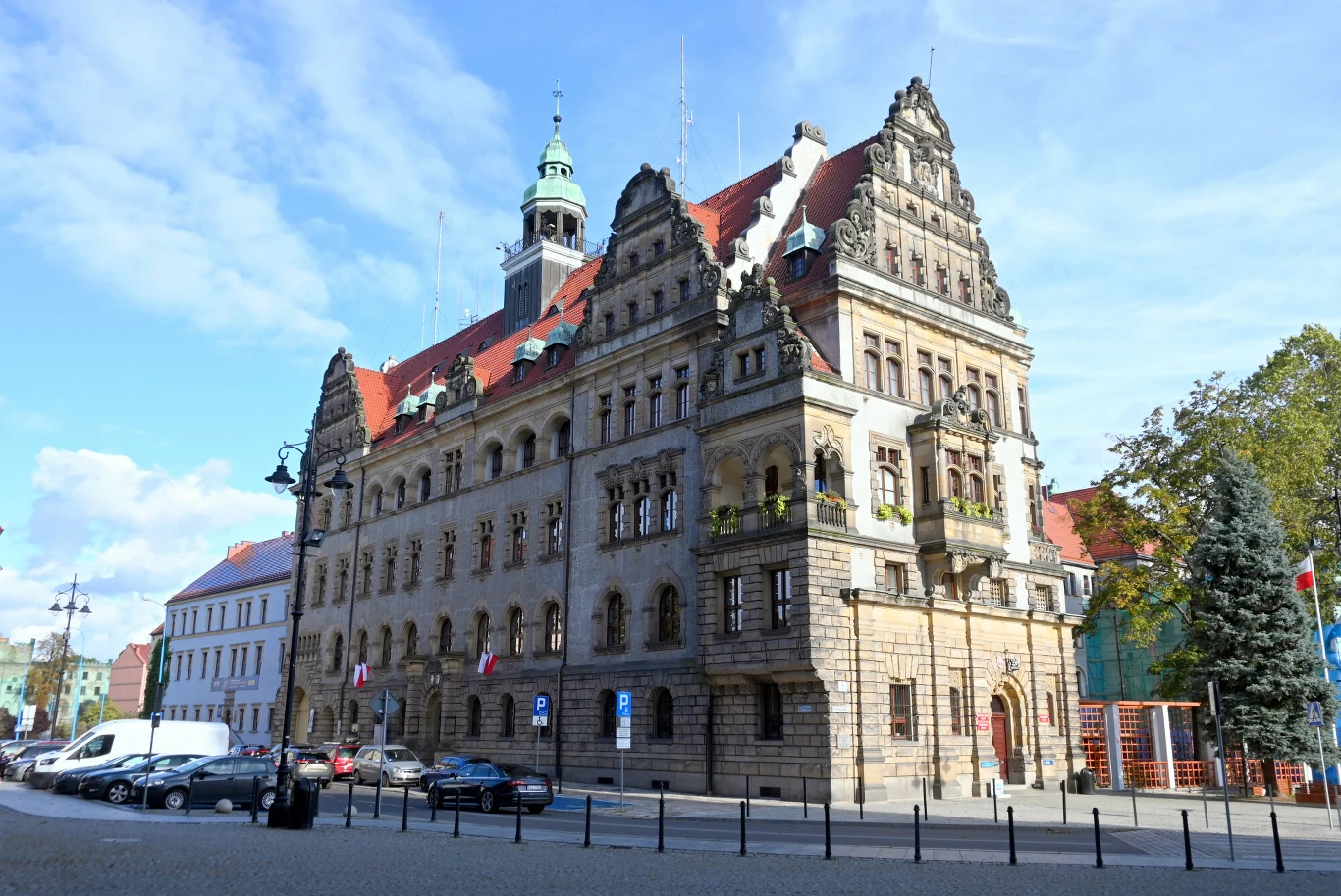 16 października mieszkańcy Legnicy mogą się zapisać na bezpłatne badania dermatoskopowe w kierunku czerniaka. Zdj. ilustracyjne Zabytkowy budynek w Legnicy z czerwonym dachem i wieżą, przed nim samochody i czarne słupki.