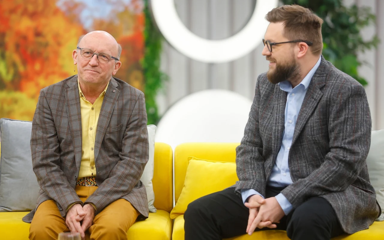 Artur Barciś z synem Franciszkiem w studiu "halo tu polsat" Dwóch mężczyzn siedzących na żółtej sofie, obaj ubrani w marynarki w kratę, rozmawiających i uśmiechniętych, na tle jesiennej dekoracji i białych geometrycznych elementów.