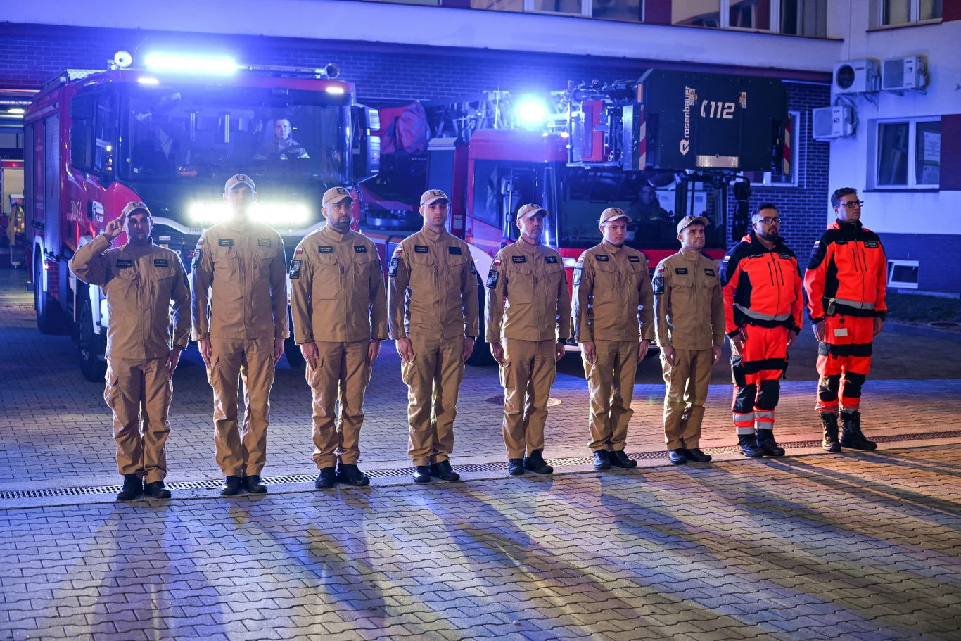 Strażacy z Komendy Miejskiej PSP w Rzeszowie uczcili pamięć dwóch kolegów, którzy zginęli podczas akcji gaśniczej w Nisku Grupa strażaków i ratowników w mundurach stoi w szeregu przed wozami strażackimi oświetlonymi niebieskimi światłami, jedna osoba salutuje.