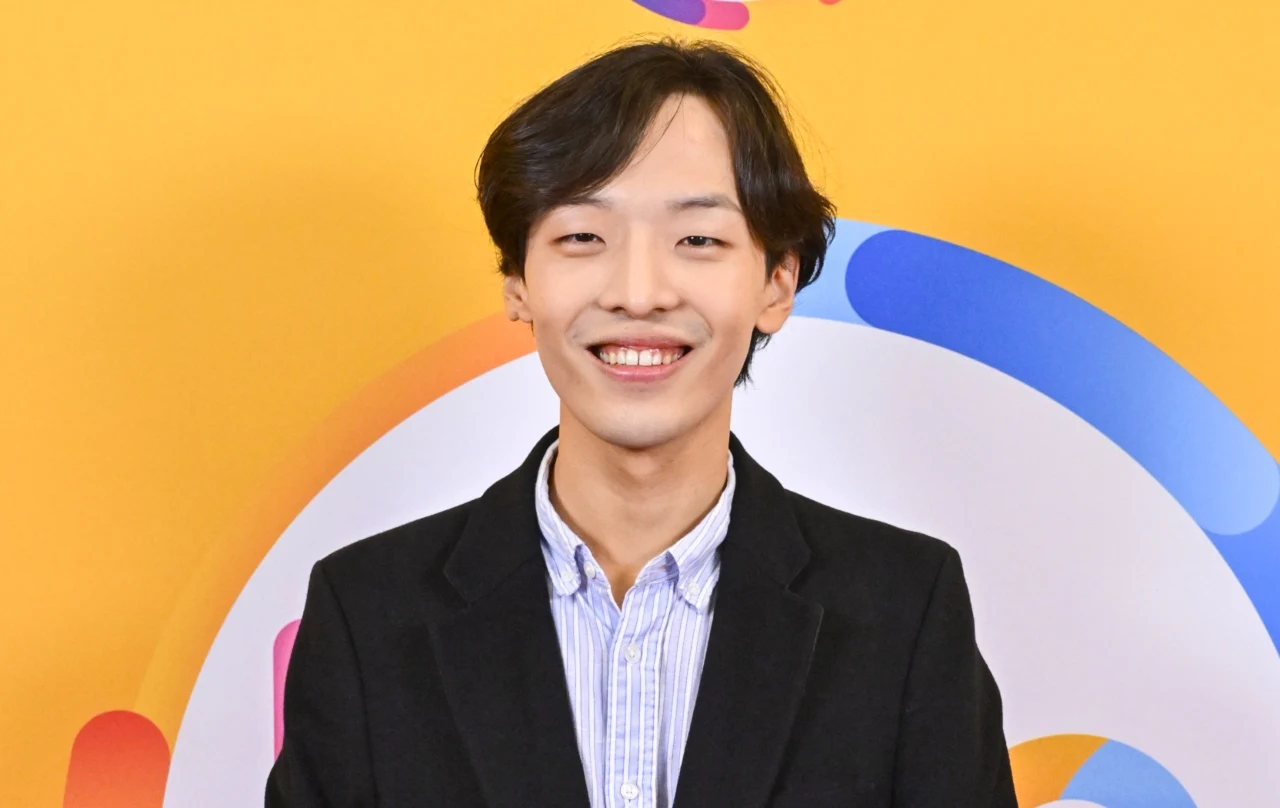 Hyuk Lee w studiu "halo tu polsat" Hyuk Lee w studiu 'halo tu polsat', w czarnej marynarce i niebiesko-białej koszuli na tle żółtej ściany.