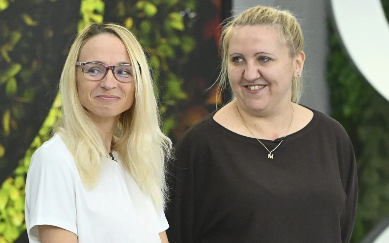 Dwie uśmiechnięte kobiety stojące obok siebie, jedna w białej bluzce i okularach, druga w czarnej bluzce z naszyjnikiem, na tle zielonej roślinności.