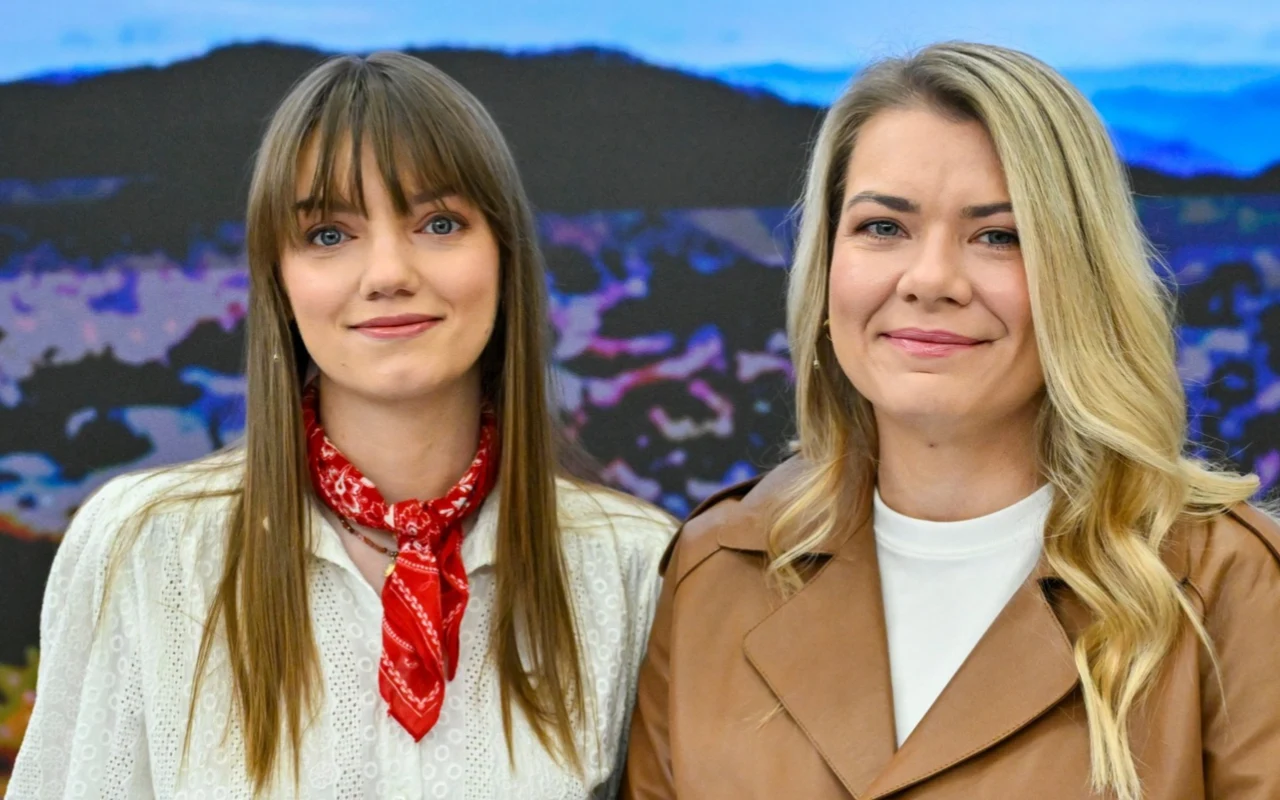 Dwie kobiety stoją obok siebie, jedna ubrana w białą bluzkę z czerwoną apaszką, druga w jasnobrązowy płaszcz i białą bluzkę, obie uśmiechają się w stronę obiektywu, w tle rozmyty krajobraz górski.