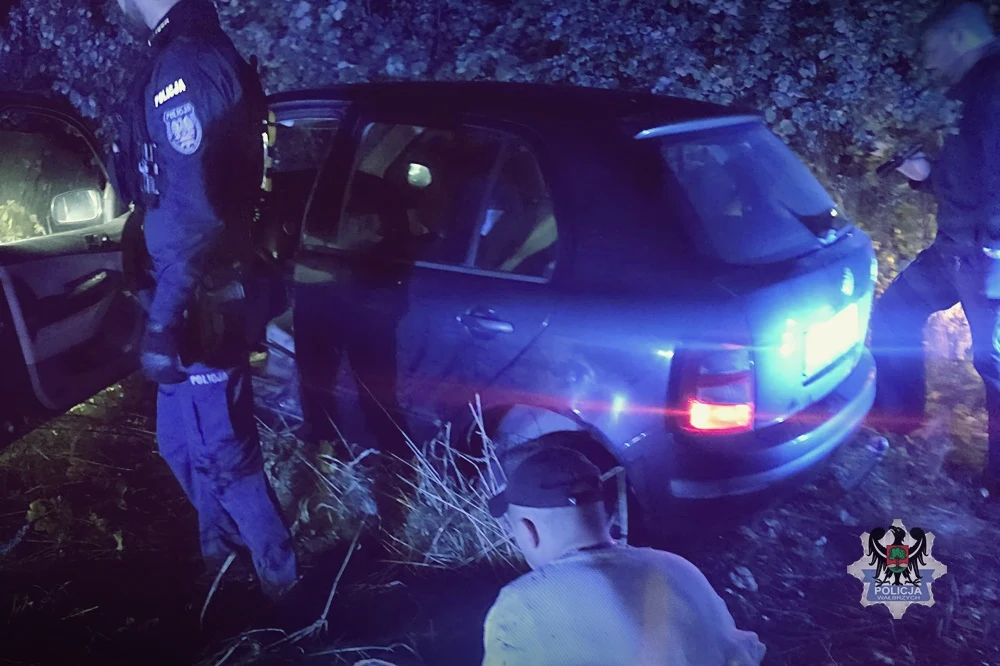 Policjanci w mundurach stojący przy zatrzymanym aucie osobowym w terenie zarośniętym roślinnością, na pierwszym planie mężczyzna w jasnej bluzie siedzący na ziemi, a w tle widoczne oznakowanie policyjne.