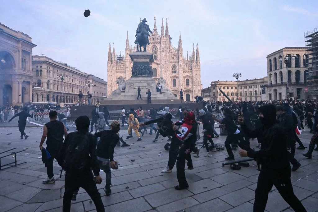 Włochy. Zamieszki na Piazza Duomo w Mediolanie Zamaskowani uczestnicy zamieszek rzucają przedmiotami na placu przed katedrą, w tle widoczny konny pomnik i gotycka fasada dużego kościoła.
