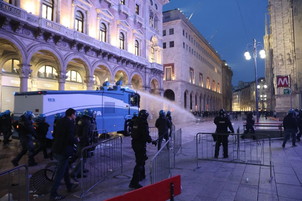 Mediolan. Policja próbowała stłumić zamieszki Oddziały policji w pełnym rynsztunku kierują armatkę wodną w stronę tłumu na miejskim placu otoczonym klasyczną architekturą.