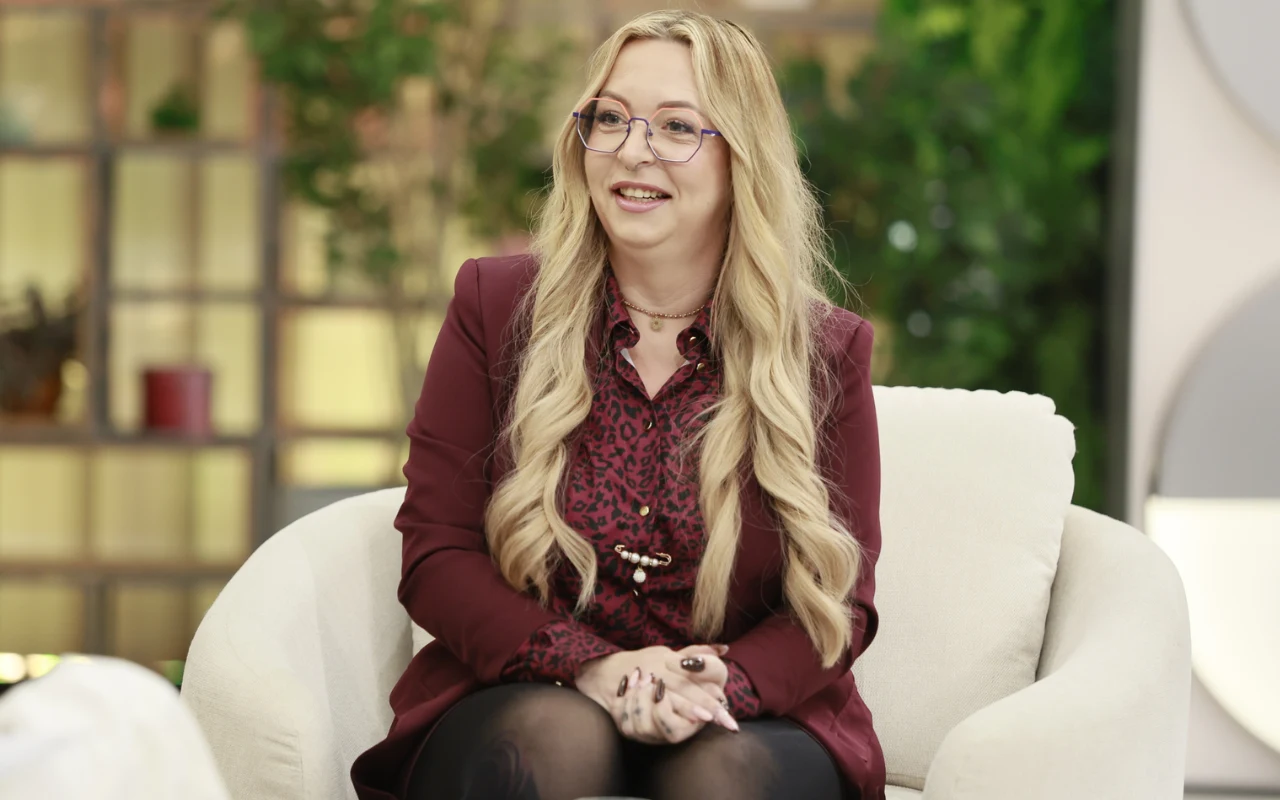 Klaudia Klimczak w "halo tu polsat" Kobieta z długimi blond włosami siedząca w jasnym fotelu, ubrana w bordową marynarkę i koszulę z wzorem, nosi okulary i uśmiecha się.