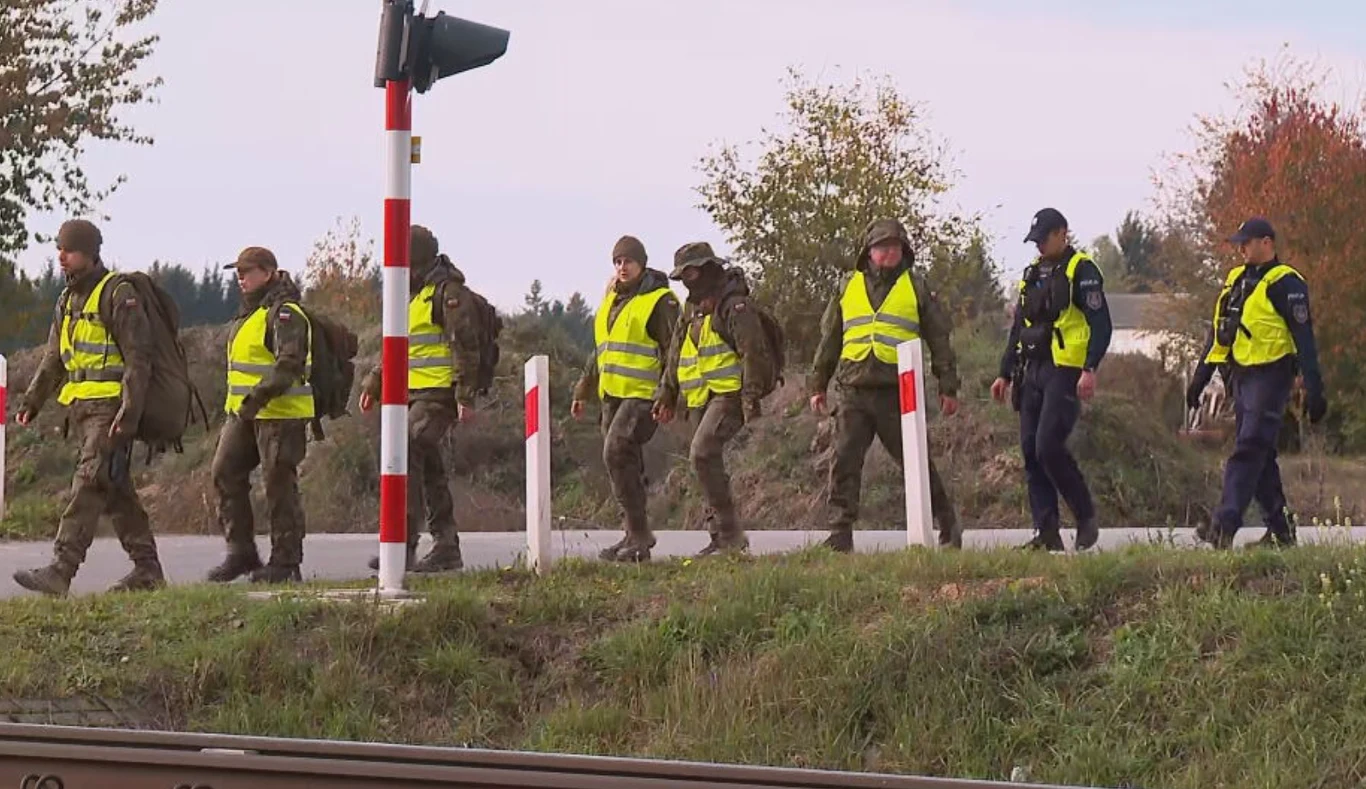 Grupa osób w mundurach i kamizelkach odblaskowych idąca wzdłuż drogi, obok funkcjonariuszy policji, z widoczną sygnalizacją kolejową i barierkami przy przejściu.