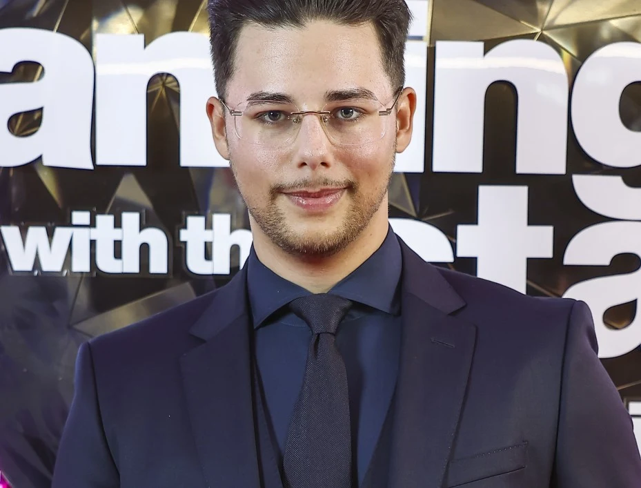 Allan Krupa Mężczyzna w eleganckim garniturze i okularach stoi przed ścianą ozdobioną dużym, białym napisem.