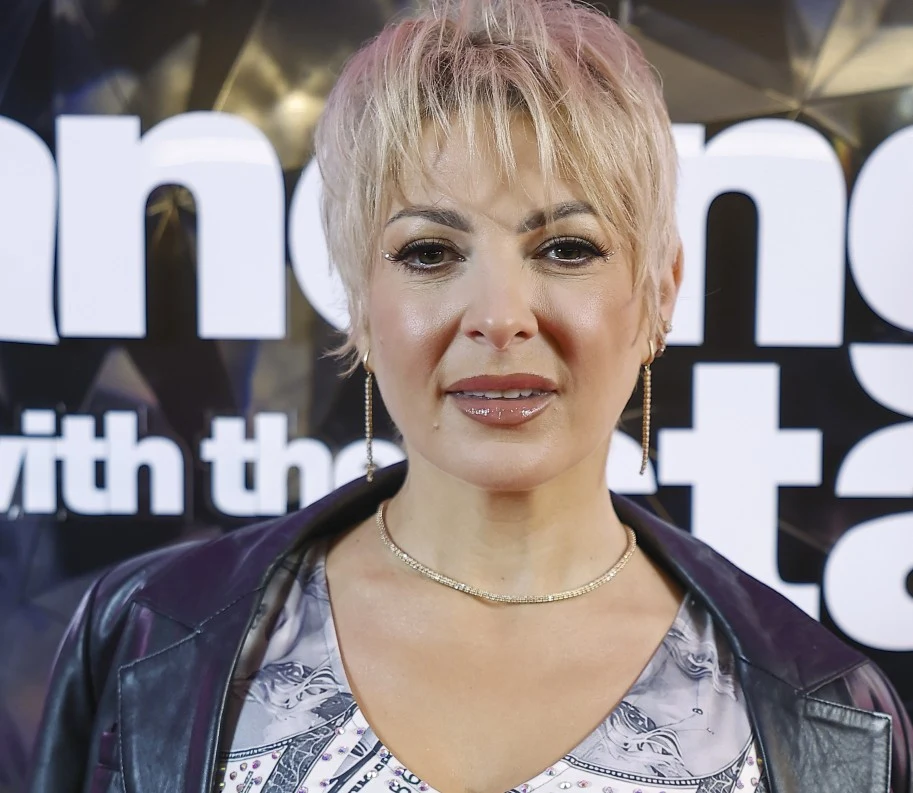 Magdalena Narożna Kobieta z krótkimi blond włosami, w lekkim makijażu, ubrana w skórzaną kurtkę i jasną bluzkę z nadrukiem, pozująca na tle napisu będącego częścią scenografii telewizyjnej.