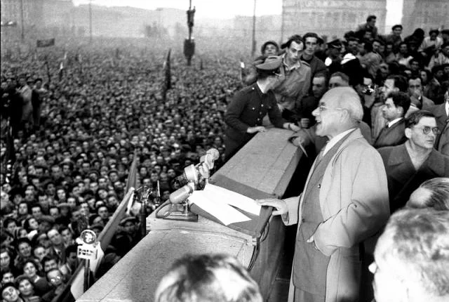 Przemówienie Władysława Gomułki na Placu Defilad, 24.10.1956r. Przywódca wygłaszający przemówienie z podwyższenia do ogromnego, szczelnie wypełnionego tłumu słuchaczy, w otoczeniu najbliższych współpracowników i przy mikrofonach.