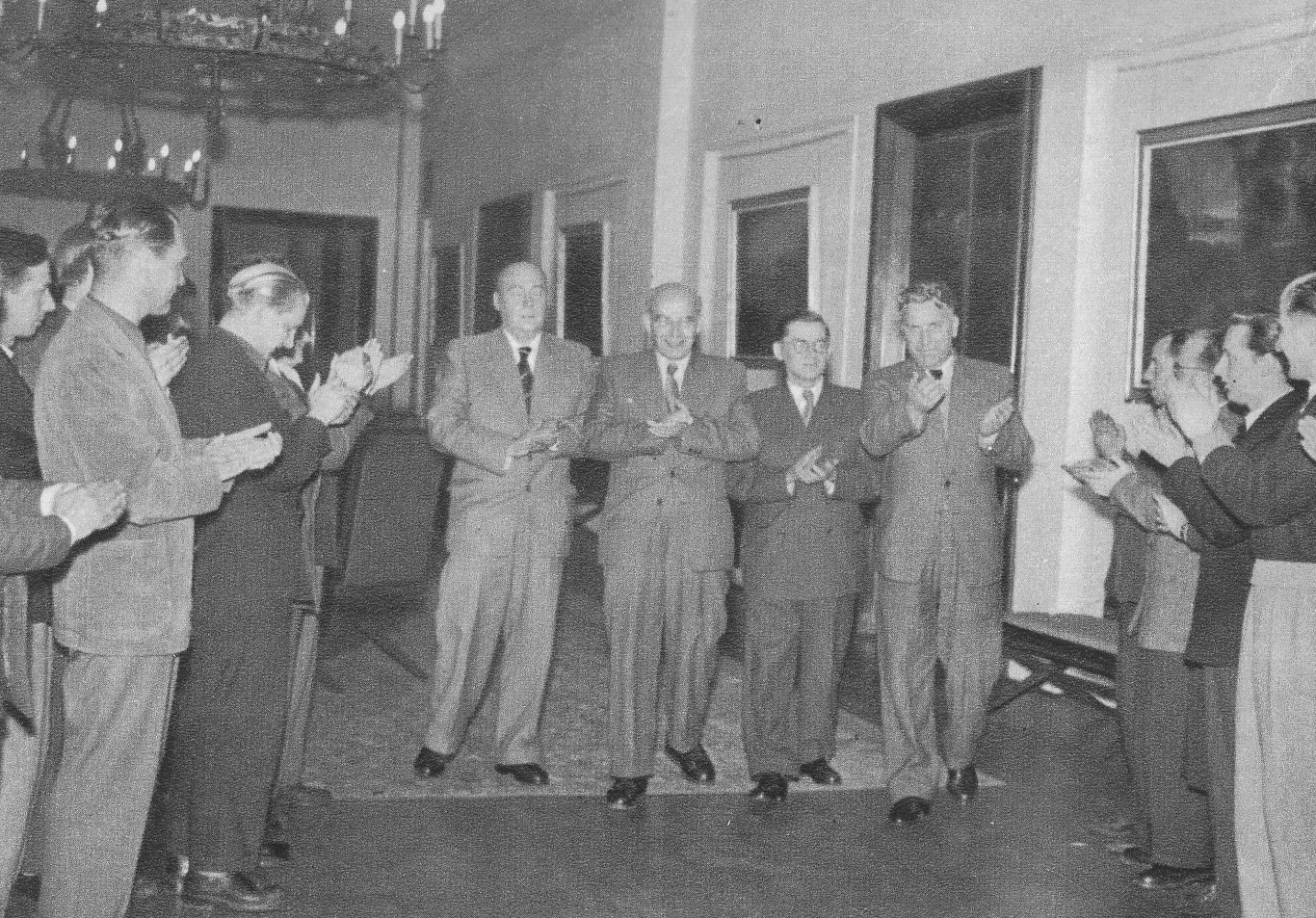 Władysław Gomułka podczas VIII Plenum Komitetu Centralnego PZPR, 10.1956r. Grupa elegancko ubranych mężczyzn ustawionych w półkolu, bijących brawo czterem mężczyznom stojącym na środku, w jasnym, przestronnym pomieszczeniu o wysokich oknach.