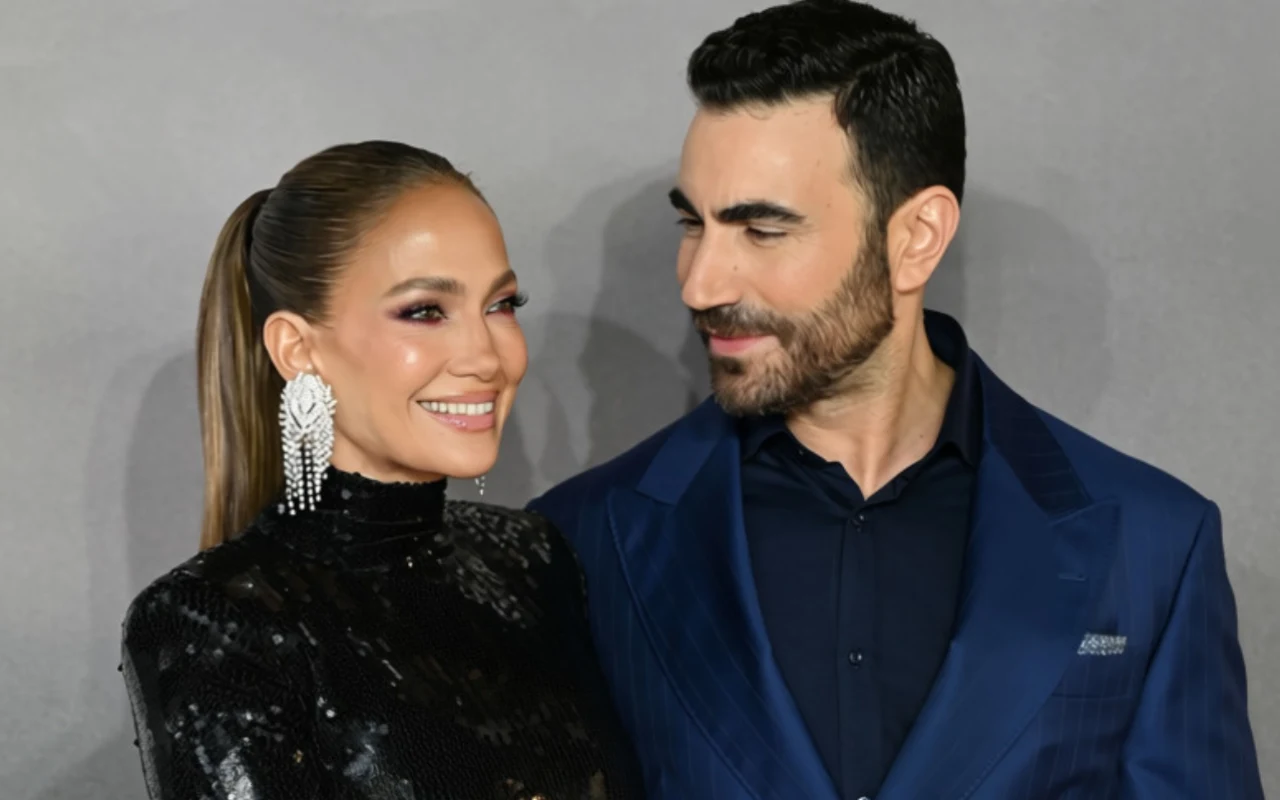 Jennifer Lopez i Brett Goldstein na premierze filmu "Biurowy romans" Uśmiechnięta kobieta w czarnej, błyszczącej sukni z efektownymi kolczykami stoi obok elegancko ubranego mężczyzny w granatowym garniturze, oboje wyglądają na zadowolonych i patrzą na siebie z sympatią.