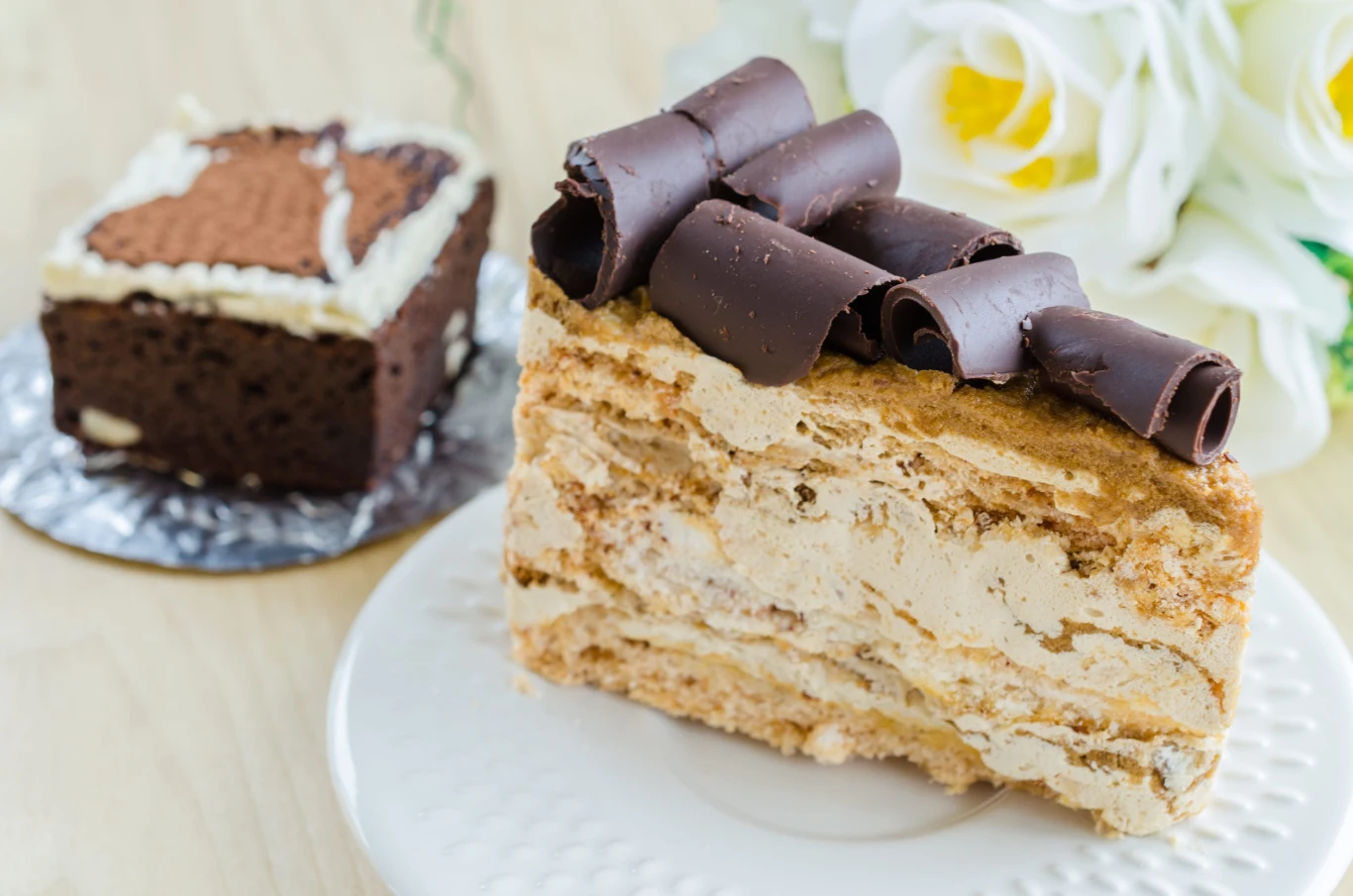 Przysmak hrabiego to połączenie kruchej bezy, czekolady, kawy i serka mascarpone w jednym deserze Kawałek bezowego ciasta z kremem mascarpone i czekoladowymi wiórami na talerzyku, w tle ciasto i kwiaty.
