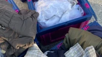 Sterta używanych ubrań, w tym koszula w kratę, kurtka i spodnie, rozłożonych przy plastikowych skrzyniach.