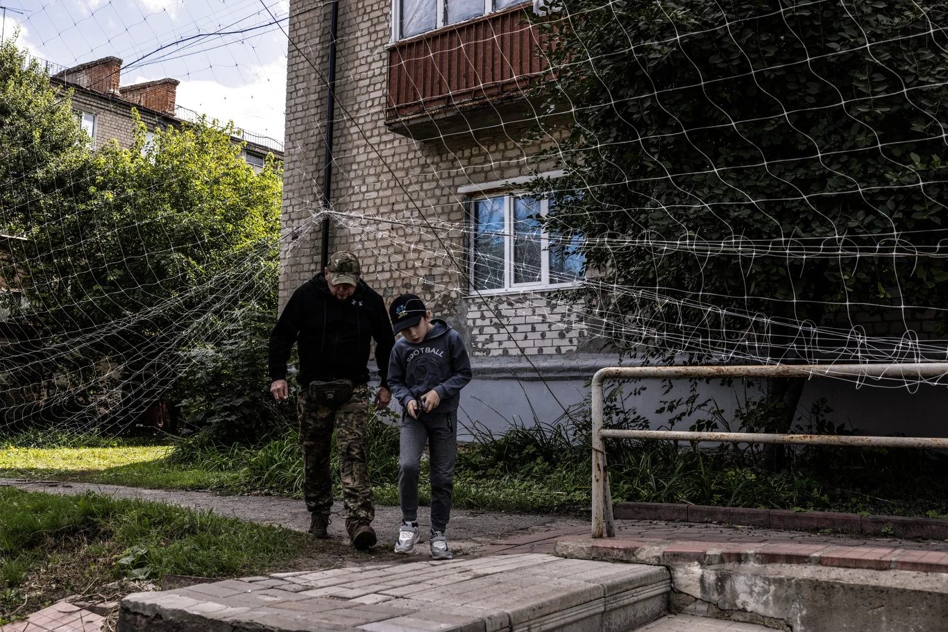 Ukraina: Siatki ochronne w Iziumie Dwie osoby, dorosły mężczyzna w ubraniu militarnym i dziecko, idą razem wzdłuż chodnika obok ogrodzonego metalową siatką budynku mieszkalnego.