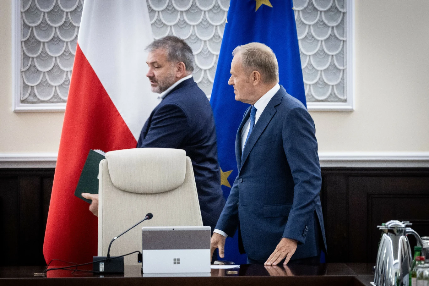 Dwaj mężczyźni w garniturach stoją obok siebie w biurowym wnętrzu, z widocznymi flagami Polski i Unii Europejskiej w tle.