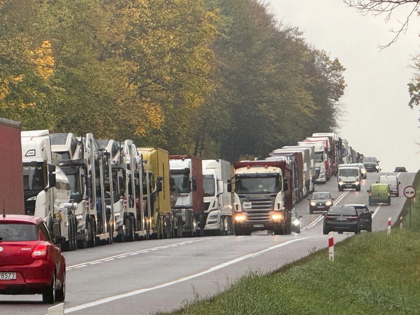 Kolejka pojazdów przed przejściem granicznym Długi korek złożony głównie z tirów i samochodów ciężarowych ustawionych jeden za drugim na dwupasmowej szosie, otoczony drzewami z jesiennymi liśćmi.