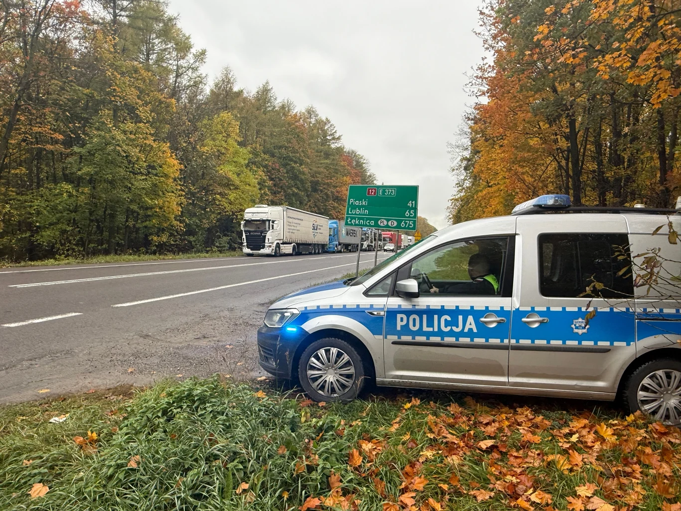 Radiowóz policyjny zaparkowany na poboczu przy drodze krajowej, w tle ciężarówka oraz zielony znak drogowy z kierunkami w gęstym, jesiennym lesie.