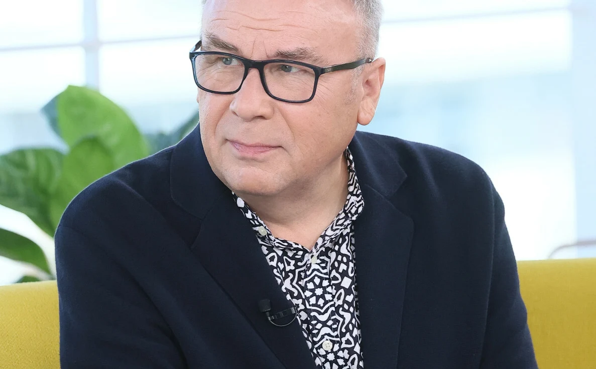 Bogdan Kalus Aktor Bogdan Kalus w ciemnej marynarce i wzorzystej koszuli siedzi na żółtej sofie.