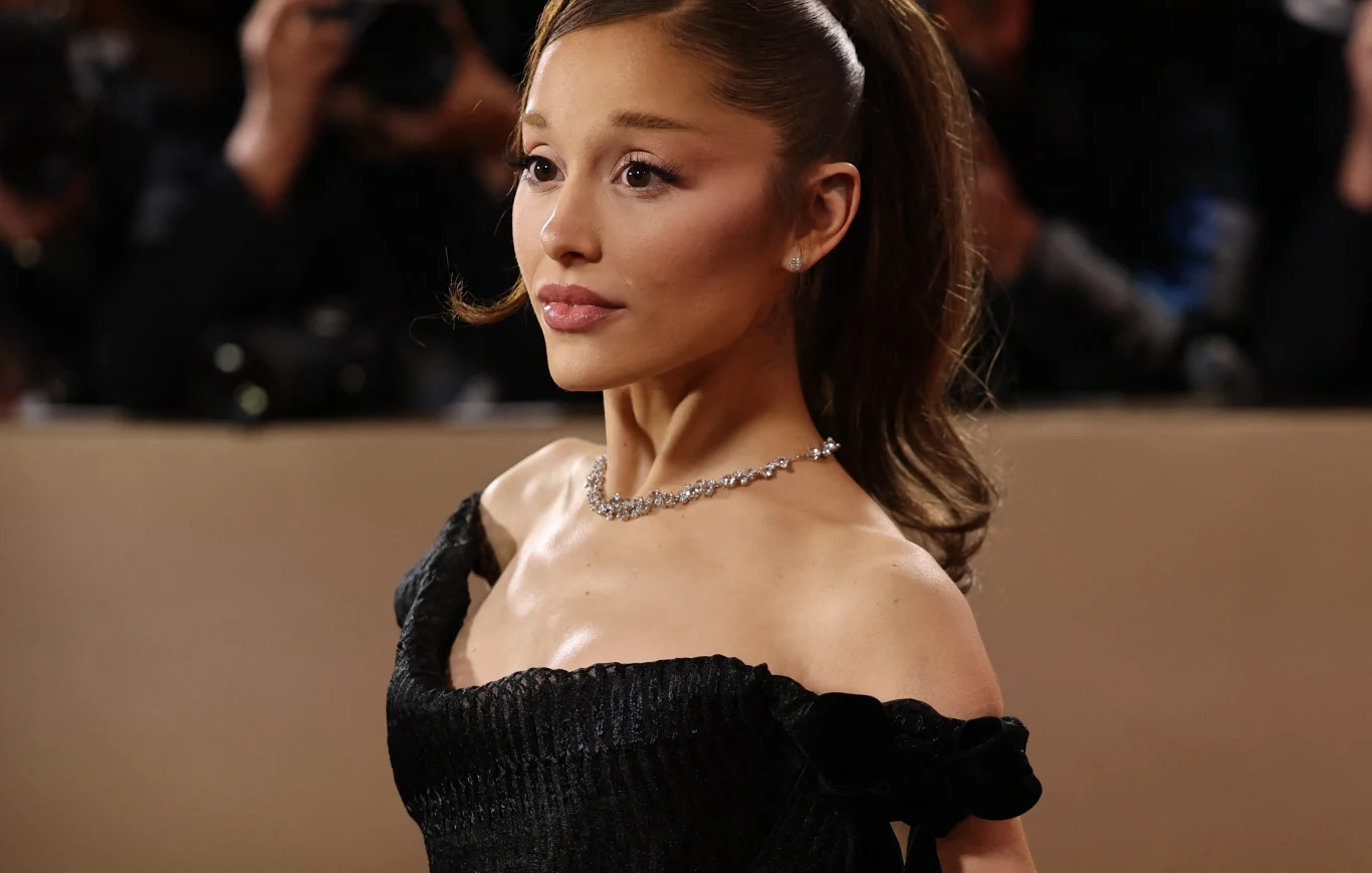 Ariana Grande Ariana Grande w eleganckiej czarnej kreacji podczas wystąpienia publicznego, w tle błyski aparatów.