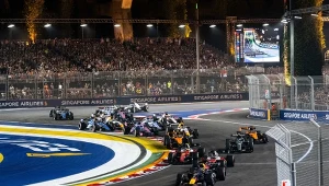 Wyścigowe bolidy pokonują zakręt toru podczas nocnego Grand Prix Formuły 1, otoczone barierami i tablicami sponsorów, a trybuny pełne są kibiców.