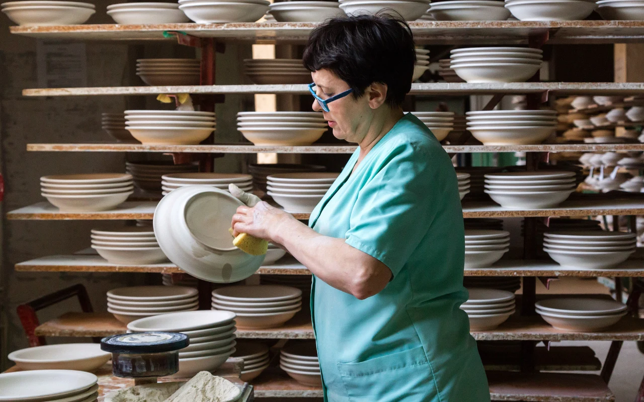 Ceramika bolesławiecka nadal powstaje dzięki pracy ludzkich rąk. Zdjęcie z zakładów ceramicznych "Bolesławiec" Kobieta w okularach i zielonym fartuchu sprawdza jakość ceramicznej miski wśród regałów wypełnionych białą porcelaną.