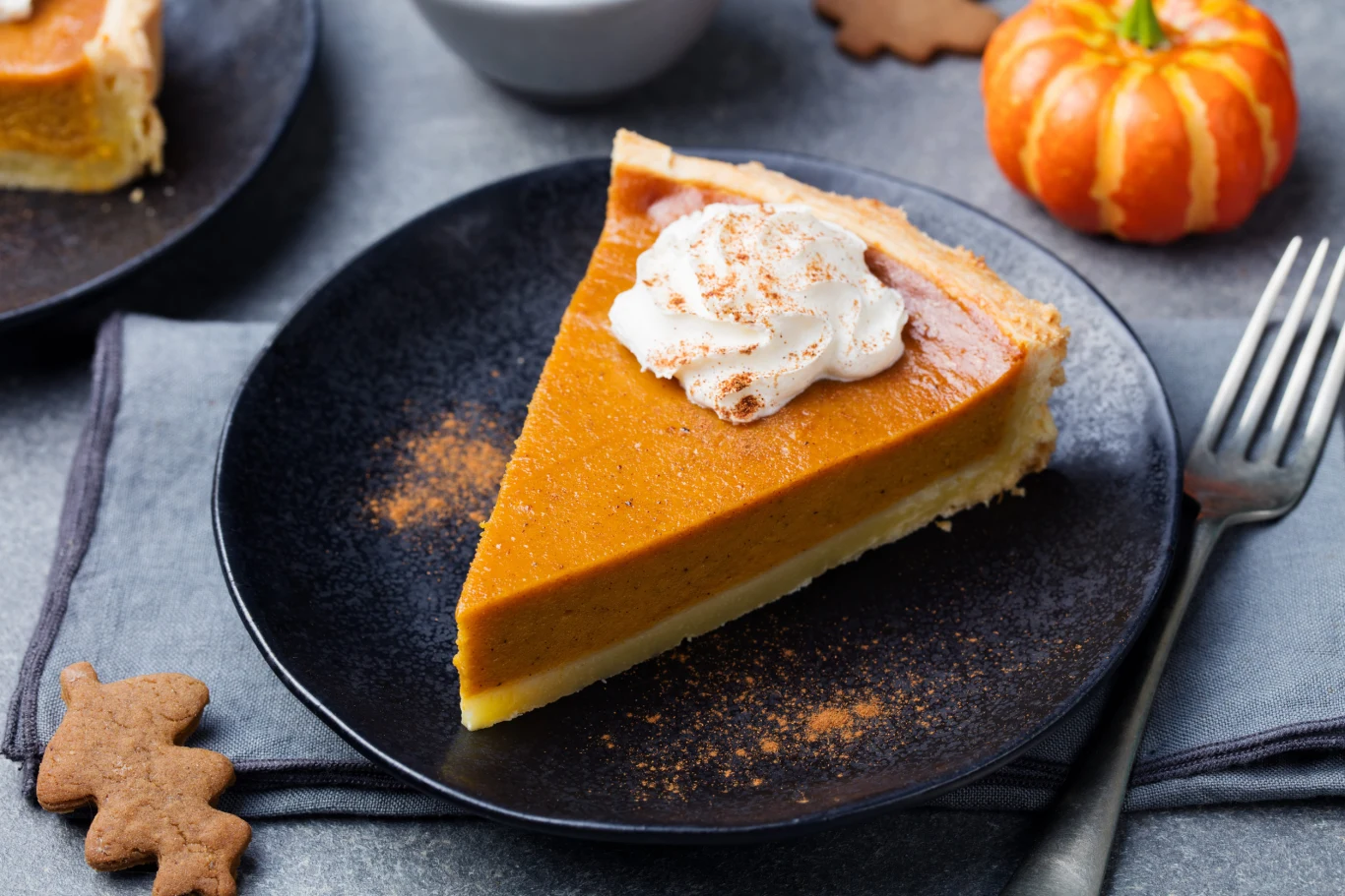 Tarta dyniowa świetnie prezentuje się podana z kleksem bitej śmietany Kawałek tarty dyniowej z bitą śmietaną i cynamonem na czarnym talerzu, obok widelca, widoczne mini dynia i pierniczki