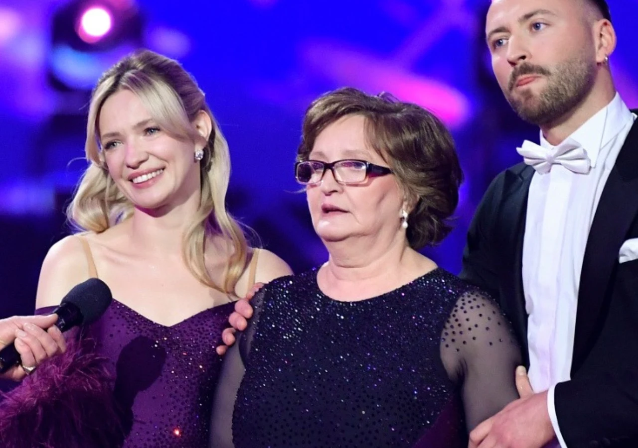 Trzy elegancko ubrane osoby stoją blisko siebie, dwie kobiety w wieczorowych sukniach w odcieniach fioletu i mężczyzna w czarnym garniturze z białą koszulą i muchą, trzymają się za ramiona, wszyscy skierowani w stronę aparatu w kolorowym oświetleniu sc...