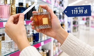 Dłonie kobiety przy ścianie z perfumami w drogerii trzymają bursztynowy flakon i papierowy pasek do testowania zapachu 