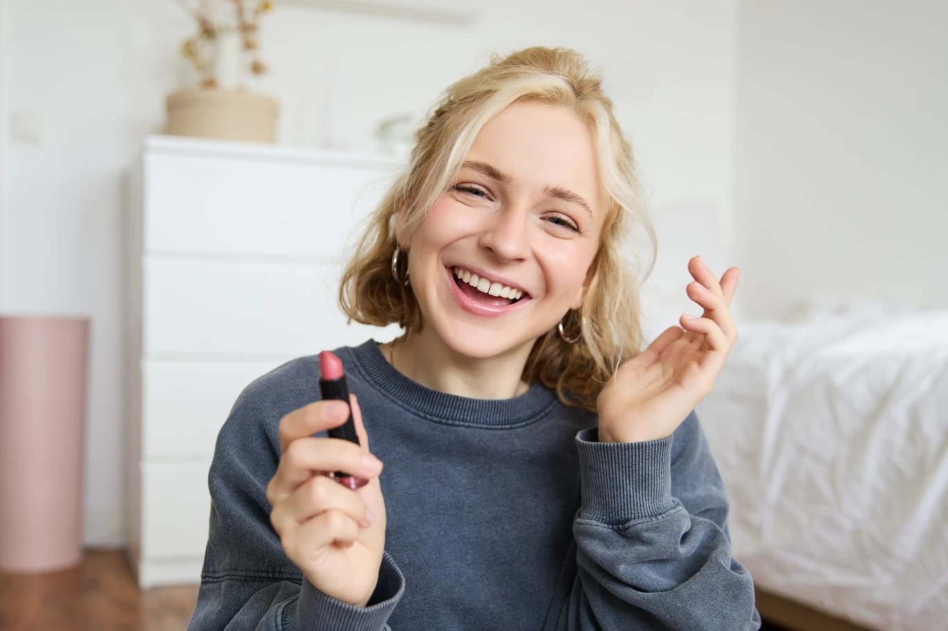 W tym sezonie schowaj maskarę do oczu i postaw na naturalny wygląd Uśmiechnięta młoda kobieta z blond włosami trzymająca szminkę w dłoni, siedząca w jasnym, przytulnym pokoju.