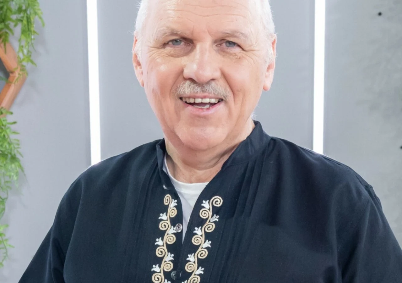 Tomasz Zubilewicz Tomasz Zubilewicz w ciemnej koszuli z haftem na tle jasnej ściany z pionowymi światłami.