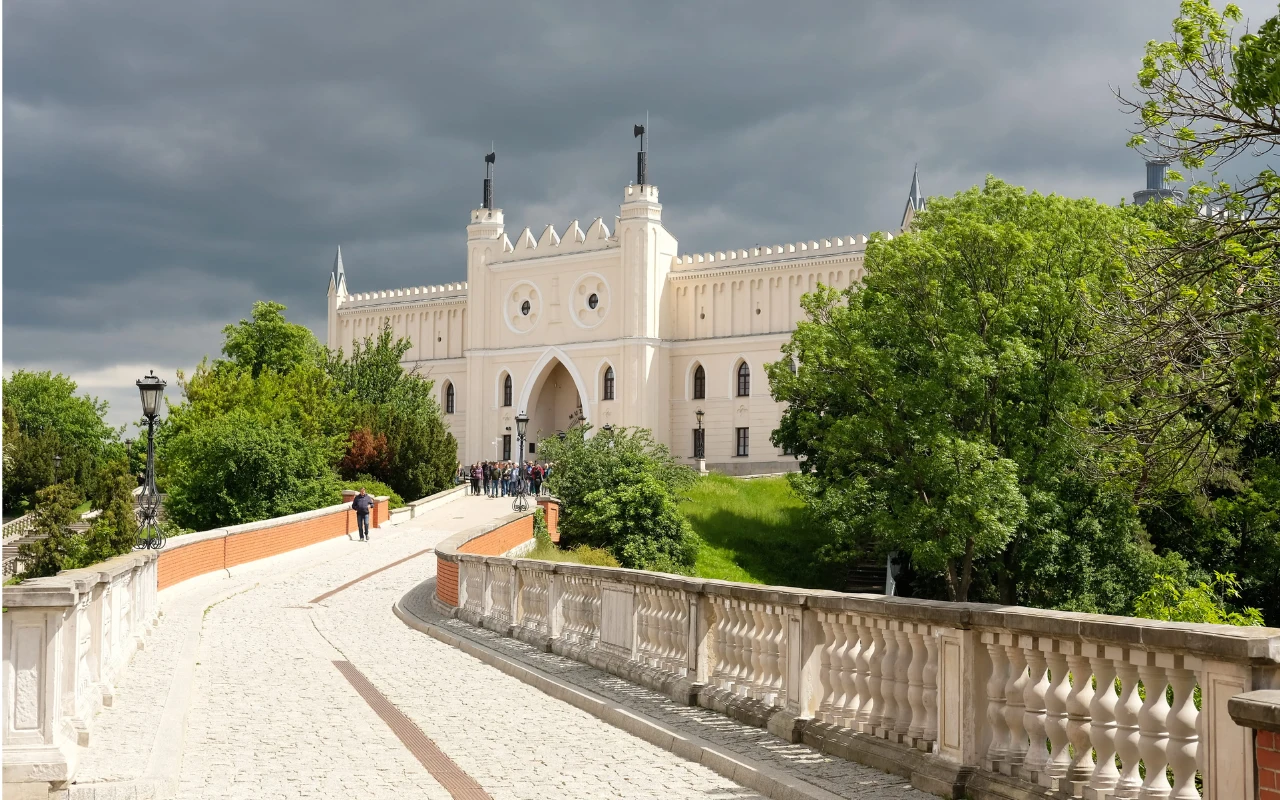 Zamek w Lublinie z charakterystyczną neogotycką wieżą bramną, połączony mostem z kamienną balustradą na tle ciemnych, burzowych chmur.