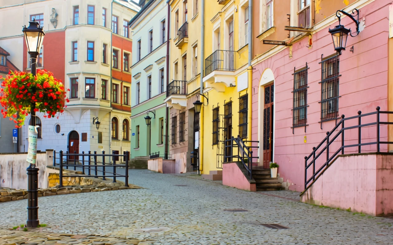 Wąska, brukowana uliczka wiodąca wzdłuż kolorowych kamienic w odcieniach różu, żółci i zieleni, ozdobiona latarniami i kwiatami.