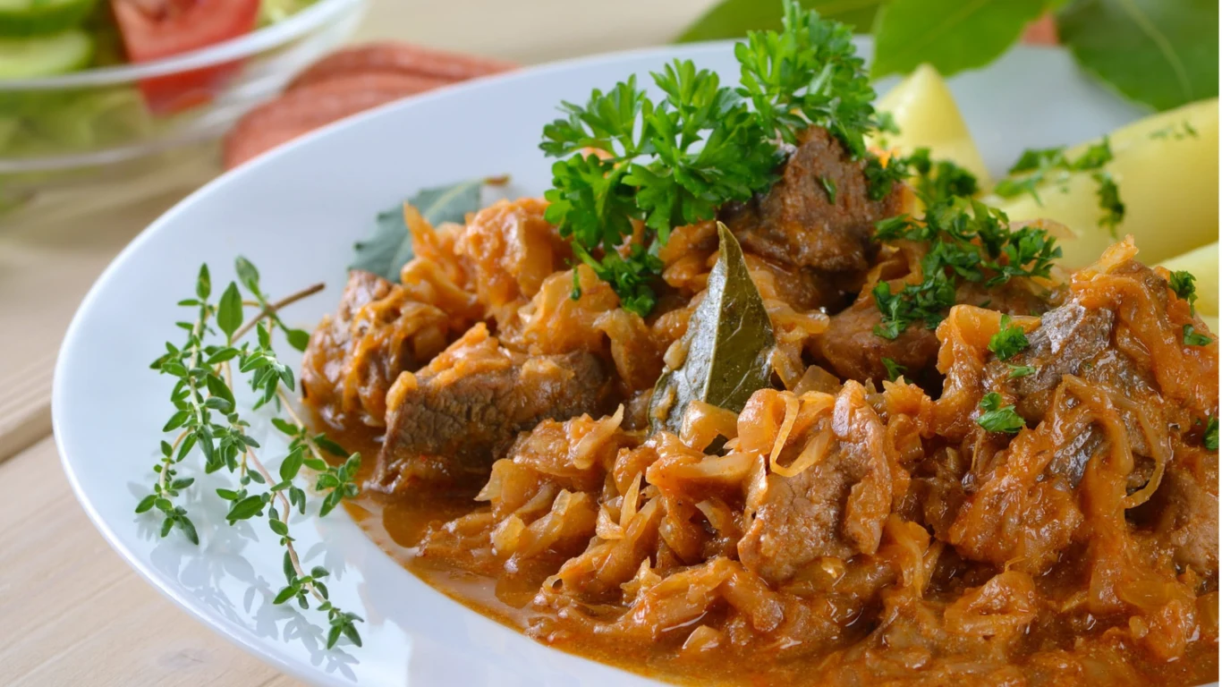 Bigos staropolski to przysmak gotowany na dwóch rodzajach kapusty z suszonymi grzybami i śliwkami Bigos staropolski z kapustą, mięsem, suszonymi grzybami i śliwkami podany na białym talerzu z natką pietruszki