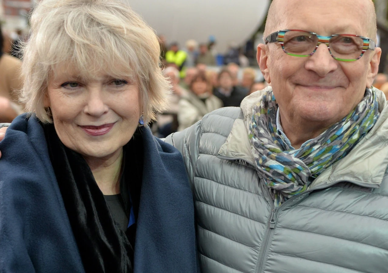 Krystyna Janda uśmiecha się, stojąc obok mężczyzny w kolorowych okularach i szaliku.