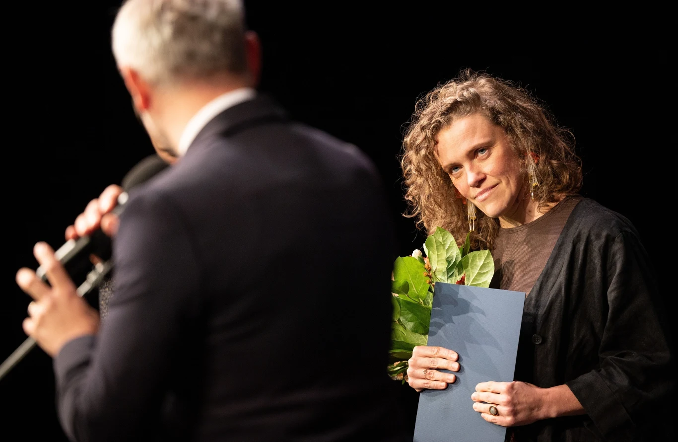 Dominika Hofman podczas uroczystości w Teatrze Śląskim Kobieta z kręconymi włosami trzyma bukiet zielonych liści i dokument, uśmiechając się lekko w stronę osoby z mikrofonem.