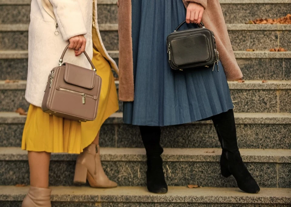 Ta spódnica pasuje do jesiennych botków! Dwie kobiety stoją na kamiennych schodach, prezentując płaszcze, spódnice oraz eleganckie torebki trzymane w rękach.