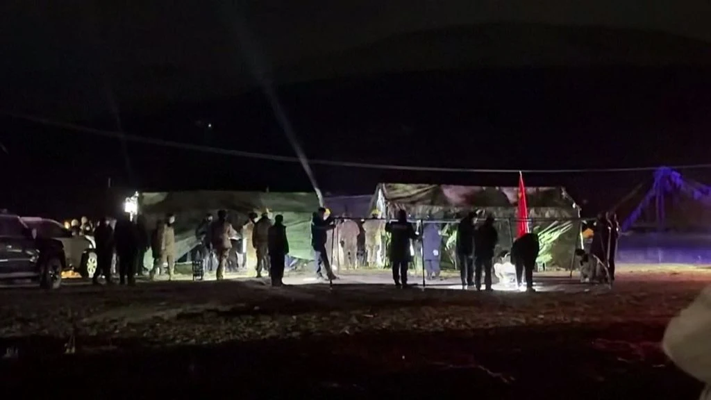 Grupa ludzi stoi nocą przy rozstawionych namiotach ratunkowych w oświetlonym terenie.