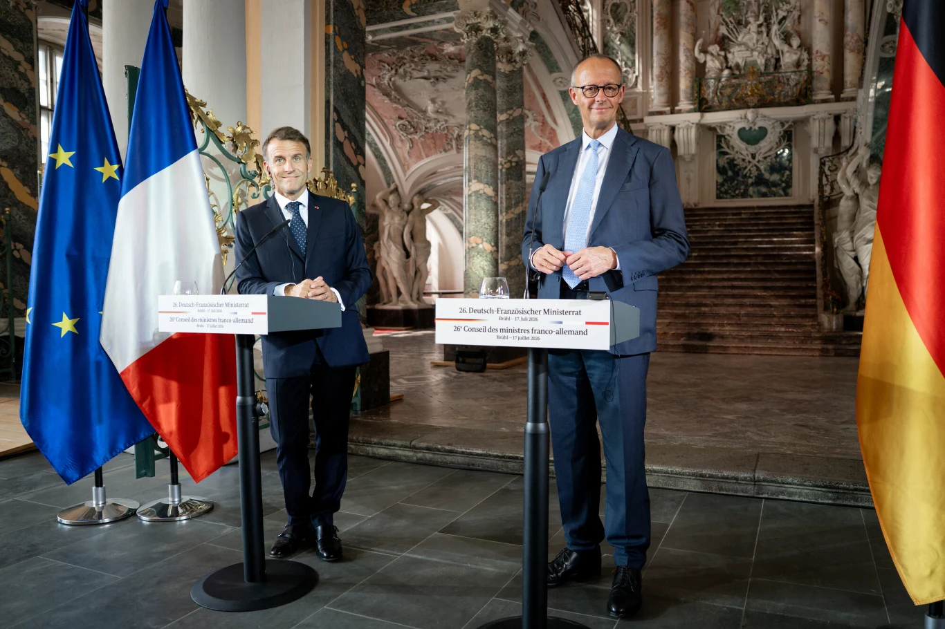 Dwóch mężczyzn w garniturach stoi za mównicami na tle dekoracyjnych schodów, po lewej widoczne flagi Unii Europejskiej i Francji, po prawej flaga Niemiec.