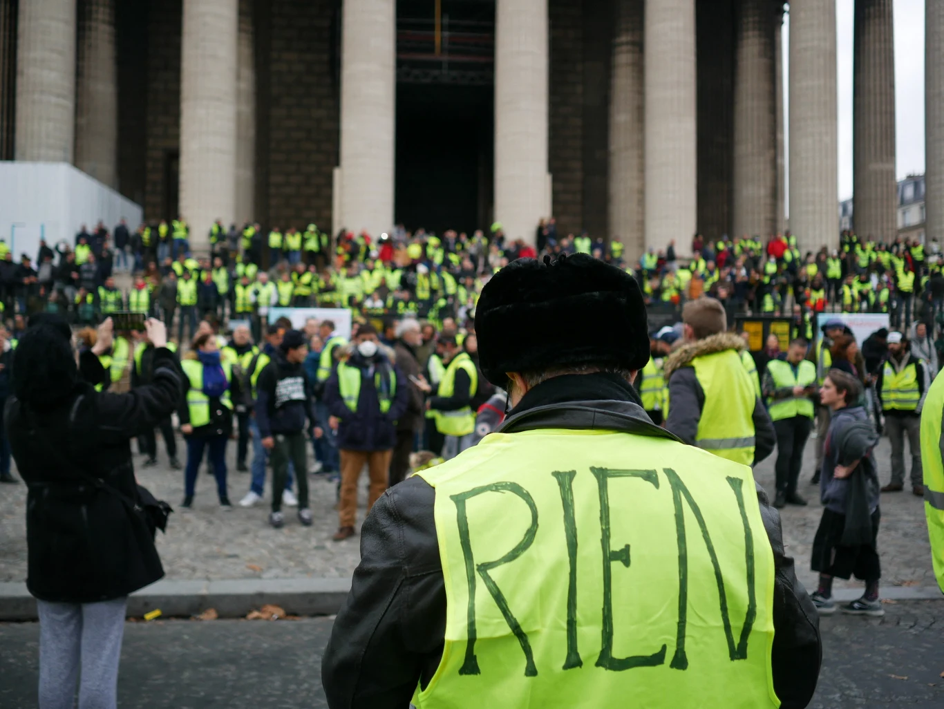 Protest "Żółtych kamizelek", listopad 2018 Tłum osób w żółtych kamizelkach zgromadzony przed monumentalnym budynkiem z kolumnami, w centrum postać w futrzanej czapce z napisem „RIEN” na plecach.