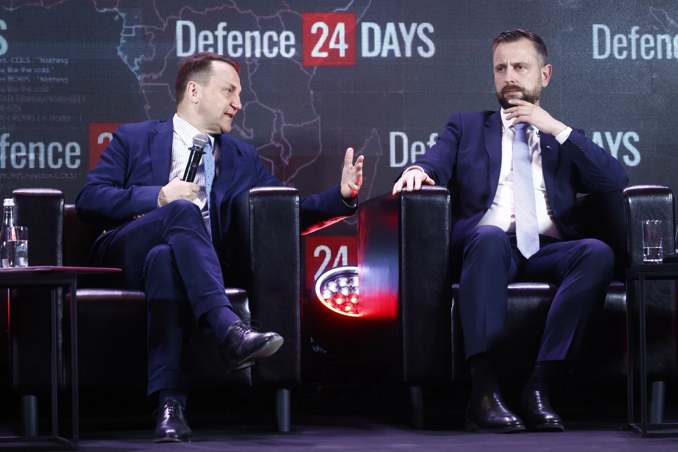 Wicepremier i szef MSZ Radosław Sikorski oraz wicepremier i szef MON Władysław Kosiniak-Kamysz Dwóch mężczyzn w garniturach siedzi w czarnych fotelach na scenie podczas konferencji Defence24 Days, prowadząc dyskusję; w tle widoczny jest ekran z nazwą wydarzenia.